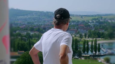 Yaşlı adam tek koluyla dikiliyor. Arka planda yeşil tepeler ve tekneler olan manzaralı bir kasabaya bakıyor.