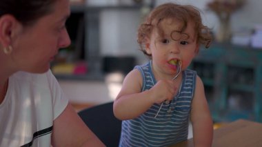 Yemek masasında çatal, dağınık yüz ve odaklanmış ifade ile makarnayı bağımsız olarak yiyen çocuk sıcak aile mutfağında ebeveynin yanında otururken