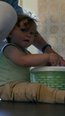 Yeni yürümeye başlayan çocuk, yanında ebeveyni olan salata fırıldağını döndürüyor, mutfak zemininde meraklı bir ifadeyle oturuyor ve kapalı bir odada aktif bir şekilde oturuyor.