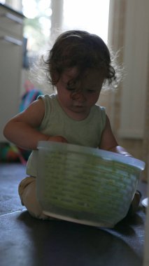 Yeni yürümeye başlayan çocuk, yerde otururken kafasına şapka gibi plastik sepet yerleştiriyor, ev ortamında mutfak eşyalarıyla yaratıcı oyunlar oynuyor.