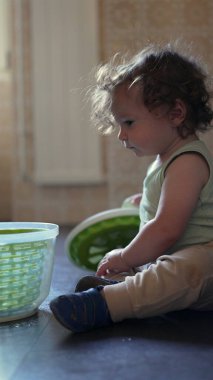 Meraklı çocuk mutfakta oturmuş plastik dönen kaseyi keşfediyor, kapağı kaldırıyor ve bağımsız oyun sırasında odaklanmış bir ifadeyle içeriye bakıyor.