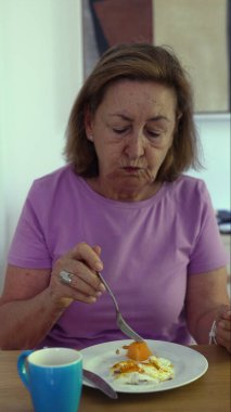 Son sınıf öğrencisi kadın kahvaltısını çırpılmış yumurta ve meyve tabağıyla yapıyor, sakin bir ev ortamında sabah ışığı eşliğinde çatal kaldırıyor.