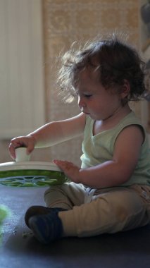 Yürüyen çocuk yerde dönen çanağı açarken tek başına mutfakta oyun oynuyordu. İçeriği merak ve ev ortamında nazik bir konsantrasyonla inceliyordu.