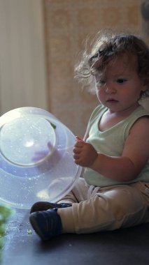 Meraklı çocuk mutfakta oturmuş plastik dönen kaseyi keşfediyor, kapağı kaldırıyor ve bağımsız oyun sırasında odaklanmış bir ifadeyle içeriye bakıyor.