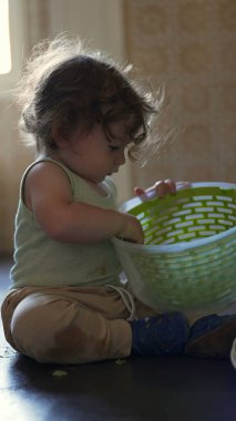 Bebek yerde otururken plastik bir çanak tutarak, tek başına oynarken ilgi ve konsantrasyonla objeyi inceliyor.