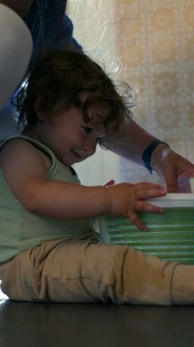 Yeni yürümeye başlayan çocuk, yanında ebeveyni olan salata fırıldağını döndürüyor, mutfak zemininde meraklı bir ifadeyle oturuyor ve kapalı bir odada aktif bir şekilde oturuyor.