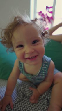 Yatakta öne eğilmiş kıvırcık saçlı, gülümseyen bebek, neşeli yüz ifadesi ve güneşli sıcak odada parlak gözleri olan pencere ve desenli battaniyeli.