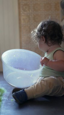 Meraklı çocuk mutfakta oturmuş plastik dönen kaseyi keşfediyor, kapağı kaldırıyor ve bağımsız oyun sırasında odaklanmış bir ifadeyle içeriye bakıyor.