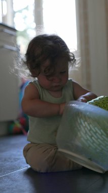 Yeni yürümeye başlayan çocuk, yerde otururken kafasına şapka gibi plastik sepet yerleştiriyor, ev ortamında mutfak eşyalarıyla yaratıcı oyunlar oynuyor.