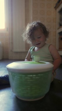 Kıvırcık saçlı çocuk, anne ve babasının yardımıyla salata kurutma makinesi çeviriyor. Sıcak ev ortamında paylaşılan mutfak etkinliği sırasında eller kapağını çevirirken odaklanmış yüz ifadesi.