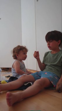 Kardeşler evde birlikte oturuyorlar, yeni yürümeye başlayan çocuk elleriyle el hareketi yapan büyük çocuğa bakıyor, ev ortamında sıradan bir aile etkileşimi.