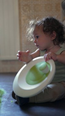 Yürüyen çocuk yerde dönen çanağı açarken tek başına mutfakta oyun oynuyordu. İçeriği merak ve ev ortamında nazik bir konsantrasyonla inceliyordu.