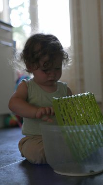 Yeni yürümeye başlayan çocuk, yerde otururken kafasına şapka gibi plastik sepet yerleştiriyor, ev ortamında mutfak eşyalarıyla yaratıcı oyunlar oynuyor.