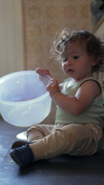 Meraklı çocuk mutfakta oturmuş plastik dönen kaseyi keşfediyor, kapağı kaldırıyor ve bağımsız oyun sırasında odaklanmış bir ifadeyle içeriye bakıyor.