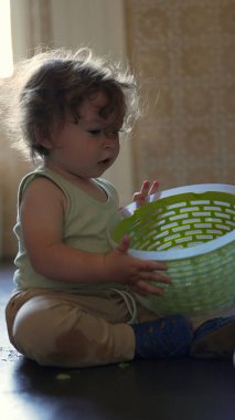 Bebek yerde otururken plastik bir çanak tutarak, tek başına oynarken ilgi ve konsantrasyonla objeyi inceliyor.