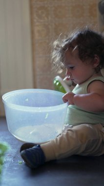 Yürüyen çocuk yerde dönen çanağı açarken tek başına mutfakta oyun oynuyordu. İçeriği merak ve ev ortamında nazik bir konsantrasyonla inceliyordu.