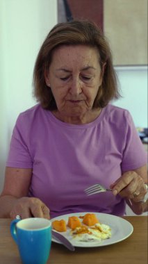 Son sınıf öğrencisi kadın kahvaltısını çırpılmış yumurta ve meyve tabağıyla yapıyor, sakin bir ev ortamında sabah ışığı eşliğinde çatal kaldırıyor.