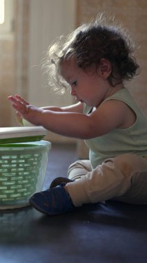 Meraklı çocuk mutfakta oturmuş plastik dönen kaseyi keşfediyor, kapağı kaldırıyor ve bağımsız oyun sırasında odaklanmış bir ifadeyle içeriye bakıyor.