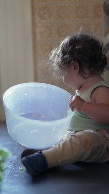 Yürüyen çocuk yerde dönen çanağı açarken tek başına mutfakta oyun oynuyordu. İçeriği merak ve ev ortamında nazik bir konsantrasyonla inceliyordu.