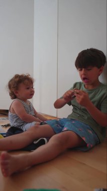 Kardeşler evde birlikte oturuyorlar, yeni yürümeye başlayan çocuk elleriyle el hareketi yapan büyük çocuğa bakıyor, ev ortamında sıradan bir aile etkileşimi.