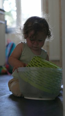 Yeni yürümeye başlayan çocuk, yerde otururken kafasına şapka gibi plastik sepet yerleştiriyor, ev ortamında mutfak eşyalarıyla yaratıcı oyunlar oynuyor.