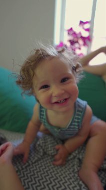 Yatakta öne eğilmiş kıvırcık saçlı, gülümseyen bebek, neşeli yüz ifadesi ve güneşli sıcak odada parlak gözleri olan pencere ve desenli battaniyeli.