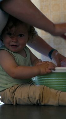 Yeni yürümeye başlayan çocuk, yanında ebeveyni olan salata fırıldağını döndürüyor, mutfak zemininde meraklı bir ifadeyle oturuyor ve kapalı bir odada aktif bir şekilde oturuyor.