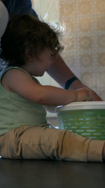 Yeni yürümeye başlayan çocuk, yanında ebeveyni olan salata fırıldağını döndürüyor, mutfak zemininde meraklı bir ifadeyle oturuyor ve kapalı bir odada aktif bir şekilde oturuyor.