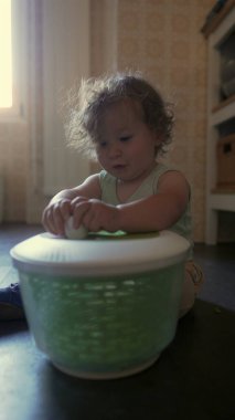 Kıvırcık saçlı çocuk, anne ve babasının yardımıyla salata kurutma makinesi çeviriyor. Sıcak ev ortamında paylaşılan mutfak etkinliği sırasında eller kapağını çevirirken odaklanmış yüz ifadesi.