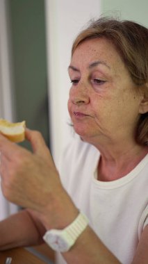 Yaşlı kadın yemek masasında bir parça ekmek ısırıyor, gevşemiş ev yemeği sırasında gözleri hafif kapalı çiğniyor.