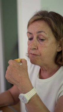 Yaşlı kadın masada otururken ekmek çiğniyor, gözleri evde sakin bir kahvaltı anında yere bakıyor.