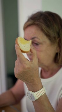 Yaşlı kadın masada otururken ekmek çiğniyor, gözleri evde sakin bir kahvaltı anında yere bakıyor.