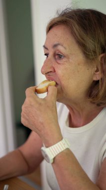 Yaşlı kadın yemek masasında bir parça ekmek ısırıyor, gevşemiş ev yemeği sırasında gözleri hafif kapalı çiğniyor.