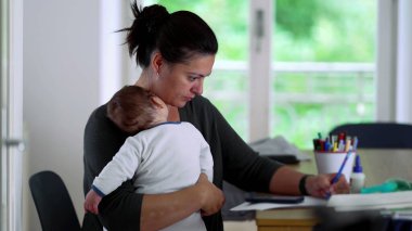 Anne, masa başında yazarken bebeğini kucağında tutuyor, işini tamamlamaya odaklanıyor, ev ortamında bakıcılık ve iş sorumluluklarını dengeliyor.