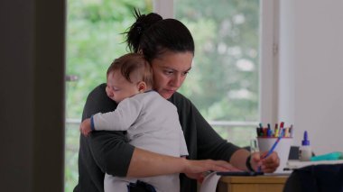 Anne, akıllı telefon kullanarak bebeğini kucağında tutuyor ve masa başında yazı yazıyor, işini dengelemeye çalışıyor ve huzurlu bir ev ortamında bakıcılık yapıyor.