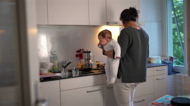 Anne mutfakta yemek hazırlarken bebeğini kucağında tutuyor, ocakta yemek pişiriyor, ev sorumluluklarını dengeliyor ve evde bakım yapıyor.