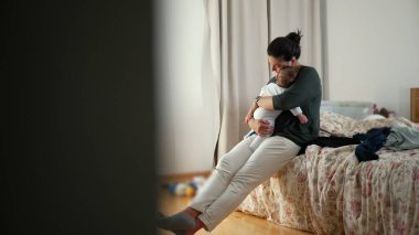 Anne yatakta oturuyor, bebeğini sakin, samimi bir şekilde kucağında tutuyor, bebeği sevgi ve özenle kucaklıyor.