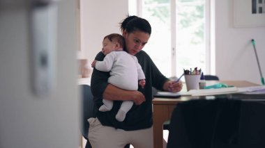 Anne bebeğini masadaki bir deftere yazarken kucağında tutuyor, çocuk bakımı ve iş arasında çoklu görev yapıyor, sakin bir ev ofisinde görevleri tamamlamaya odaklanıyor.
