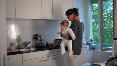 Anne mutfakta yemek hazırlarken bebeğini kucağında tutuyor, ocakta yemek pişiriyor, ev sorumluluklarını dengeliyor ve evde bakım yapıyor.