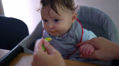 Anne bebeğe nazikçe yemek veriyor, bebeğin annesinin elini tutarken yakın plan, isteksiz yüz ifadesi, ilk kez katı gıda deneyimi, sabır ve bakım, besleyici çevre.