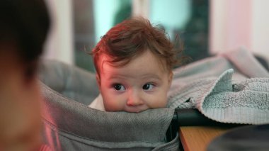 Meraklı yüz ifadesine sahip bebeğe yakın çekim, yüksek sandalyede dinlenme, nazik bakış ve rahat duruş, çevreyi gözlemleme, yumuşak ve özenli, bebeğin yüzüne odaklanma