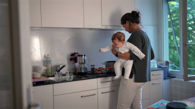Anne mutfakta yemek hazırlarken bebeğini kucağında tutuyor, ocakta yemek pişiriyor, ev sorumluluklarını dengeliyor ve evde bakım yapıyor.