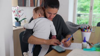 Anne, akıllı telefon kullanarak bebeğini kucağında tutuyor ve masa başında notlar alıyor. Bakıcılık görevini tamamlamaya odaklanırken aynı zamanda ev idaresini de dengeliyor.