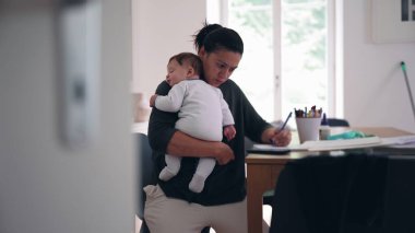 Anne bebeğini masadaki bir deftere yazarken kucağında tutuyor, çocuk bakımı ve iş arasında çoklu görev yapıyor, sakin bir ev ofisinde görevleri tamamlamaya odaklanıyor.