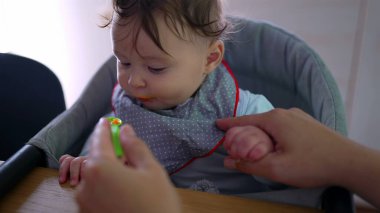 Anne bebeğe nazikçe yemek veriyor, bebeğin annesinin elini tutarken yakın plan, isteksiz yüz ifadesi, ilk kez katı gıda deneyimi, sabır ve bakım, besleyici çevre.