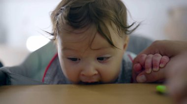 Bebek katı yiyeceği denemekte tereddüt ediyor, isteksiz ifadeye yakın çekim, ağız kenarında kaşık, anne rehberliği, ilk kez yemek yeme, ev ortamında nazik bir giriş.