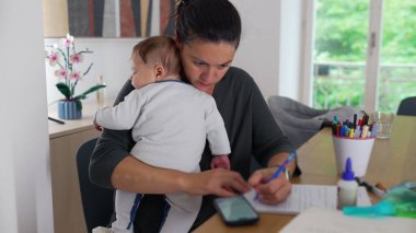 Anne, akıllı telefon kullanarak bebeğini kucağında tutuyor ve masa başında notlar alıyor. Bakıcılık görevini tamamlamaya odaklanırken aynı zamanda ev idaresini de dengeliyor.
