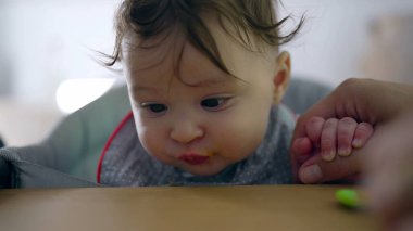Bebek katı yiyeceği denemekte tereddüt ediyor, isteksiz ifadeye yakın çekim, ağız kenarında kaşık, anne rehberliği, ilk kez yemek yeme, ev ortamında nazik bir giriş.