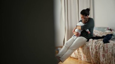Anne yatakta oturuyor, bebeğini sakin, samimi bir şekilde kucağında tutuyor, bebeği sevgi ve özenle kucaklıyor.