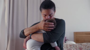 Anne bebeğinin başını öperken, huzurlu bir yuvada bir anlık şefkat ve şefkat içinde bebeğini kucaklar.